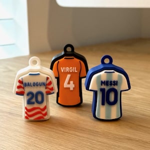 May include: Three small jersey-shaped charms with loops at the top. The left charm is white with red wavy stripes, the name BALOGUN, and number 20. The center charm is orange with black trim, the name VIRGIL, and number 4. The right charm is blue and white striped with the name MESSI and number 10.