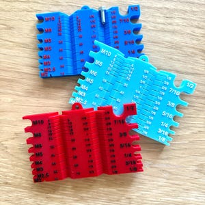 May include: Three plastic thread pitch gauges in blue and red. Each gauge has multiple teeth and measurement markings for different thread sizes, including M2.5 to M10 and fractional inch sizes. The gauges are designed for measuring screw threads.