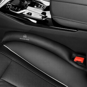 Puede incluir: Relleno negro para huecos de asiento de coche con el logotipo de Mercedes-Benz. El relleno está hecho de un material liso y curvado y se coloca entre el asiento del coche y la consola central. Se ve una hebilla roja del cinturón de seguridad.