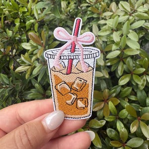 May include: Embroidered patch featuring an iced coffee drink in a clear plastic cup. The patch has a red straw and a pink bow. The drink is filled with ice cubes and a light brown liquid. The patch is surrounded by a white border.