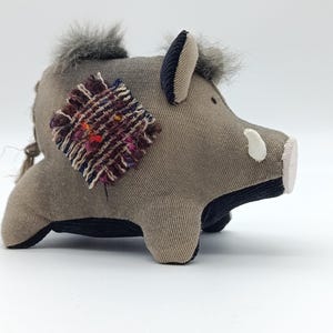 Può includere: Un giocattolo a forma di cinghiale in peluche, nei toni del grigio e del nero. L'animale di peluche ha un corpo strutturato, un ciuffo di pelo grigio e una toppa decorativa sul lato. Il muso del cinghiale è bianco e le sue orecchie sono di un colore a contrasto.