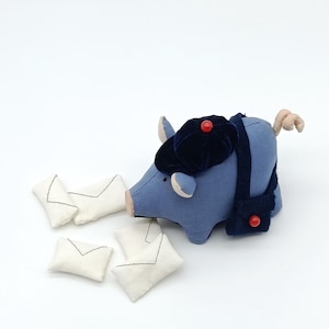 Op de afbeelding: Een blauw varkensknuffel met een donkerblauwe hoed met een rode knoop en een bijpassende tas. Verschillende kleine witte stoffen enveloppen met zwarte stiksels liggen verspreid rond het varken.