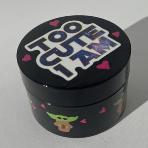Pode incluir: Um recipiente redondo preto com o texto "TOO CUTE I AM" em branco e um design de galáxia. O recipiente apresenta corações rosa e uma personagem de desenho animado. O recipiente é provavelmente usado para armazenamento.