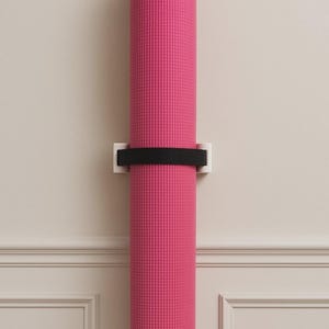 Op de afbeelding: Een opgerolde roze yogamat, vastgezet met een zwarte band en een witte wandhouder. De mat is verticaal aan een muur met decoratieve lijsten bevestigd, wat wijst op fitness of wellness.