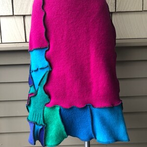 Op de afbeelding: Een levendige, knielange rok in een gedurfde fuchsia-tint, met een patchworkontwerp van turquoise, blauwe en groene viltwol. De rok heeft een geschulpte rand en een geribbelde tailleband, wat een unieke en kleurrijke stijl biedt.