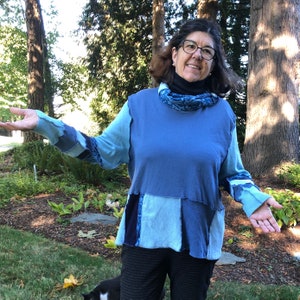 May include: A patchwork blue long-sleeve top with various shades of blue fabric. The top has a round neckline and is worn with a black turtleneck and patterned scarf. The person is wearing glasses and dark pants.
