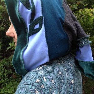 Könnte beinhalten: Eine Patchwork-Kapuze in Blau-, Türkis- und Grautönen mit grünen Blattanwendungen. Die Kapuze ist aus verschiedenen Stoffen gefertigt, darunter Samt und Fleece. Die Person trägt ein Oberteil mit türkisfarbenem und blauem Muster.