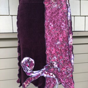 May include: A unique, handmade skirt featuring a combination of textures and patterns. The skirt is primarily a deep purple, with a velvet-like fabric on one side and a floral lace design on the other. Decorative fabric accents add a whimsical touch.