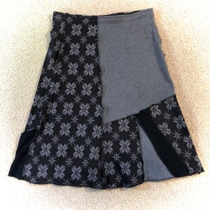 May include: A-line skirt with a patchwork design. The skirt features a combination of black fabric with a gray snowflake pattern and solid gray fabric. The skirt has a slit on the front and a black waistband.