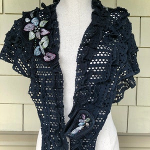 May include: A black crocheted shawl with ruffled edges and floral embroidery. The shawl is displayed on a white mannequin.
