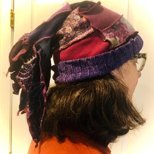 Pode incluir: Um gorro de tricô roxo, vermelho e verde com uma longa cauda roxa que flui. O gorro é feito de uma variedade de tecidos, incluindo lã, veludo e algodão.