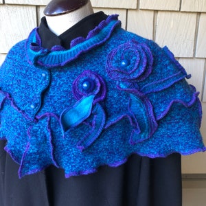 May include: A blue and purple knitted shawl with a ruffled edge and decorative flower embellishments. The shawl is made of a soft, textured yarn and features a unique, hand-crafted design.