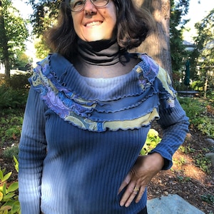May include: A blue long-sleeved shirt with a ruffled neckline and a floral pattern. The shirt is made of a soft, ribbed fabric.