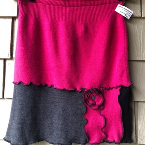 May include: A two-tone skirt in shades of hot pink and charcoal grey. The top portion is a solid hot pink, while the bottom is grey with a decorative pink panel and a flower accent. The skirt is displayed on a black hanger.