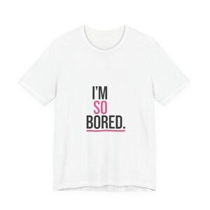Puede incluir: Camiseta blanca con el texto "I'M SO BORED." en negro y rosa. El texto está centrado en la parte delantera de la camiseta. La camiseta tiene mangas cortas y cuello redondo.