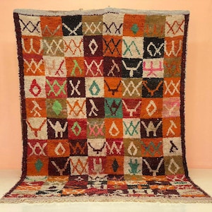 May include: A vibrant, patterned rug with a geometric design. The rug displays a patchwork of squares in orange, brown, black, and cream. It features various shapes and symbols, woven from a textured material with a fringed border.
