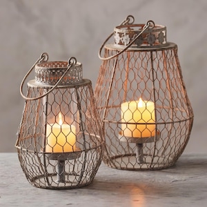Può includere: Due lanterne rustiche in metallo con un design a rete metallica. Ogni lanterna ha un manico e una candela all'interno, creando un bagliore caldo. Le lanterne sono di dimensioni variabili e sono realizzate in metallo color bronzo.