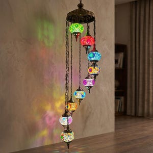 May include: A multi-colored, hanging pendant light fixture with mosaic glass globes. The fixture has a bronze-colored metal frame and chain. The globes are in various colors, including green, red, blue, yellow, and purple. The light casts colorful patterns on the wall.