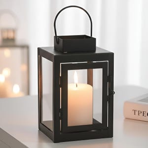 Può includere: Una lanterna in metallo nero con una cornice quadrata e pannelli di vetro trasparente. All'interno brucia una candela bianca, che emana un bagliore caldo. La lanterna ha un manico e un piccolo vassoio sulla parte superiore. Il design è moderno e minimalista.