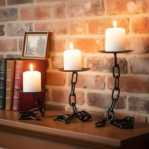 May include: Three black metal candle holders with white pillar candles. The candle holders are made of chain links and have a round base and a round tray to hold the candle. The background is a brick wall.