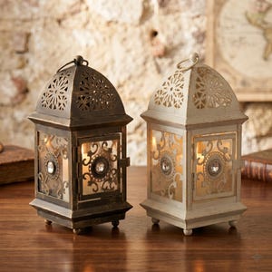May include: Two decorative lanterns. One is bronze-colored, the other is off-white. Both have intricate cut-out designs on the top and sides, with glass panels and a central jewel. They are designed to hold candles, creating a warm, inviting glow.