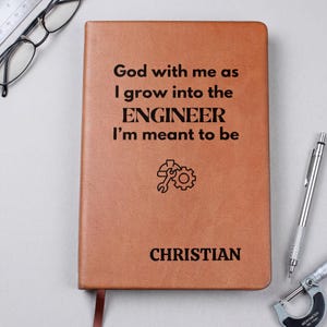 May include: A tan notebook with black text that reads God with me as I grow into the ENGINEER I'm meant to be, followed by a graphic of a hard hat and gear, and the name CHRISTIAN at the bottom. The notebook is rectangular with rounded corners.