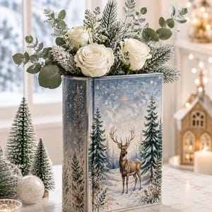 Può includere: Un vaso in ceramica rettangolare a forma di libro, decorato con una scena di bosco invernale con un cervo, alberi di pino e una stella. Il vaso è riempito con rose bianche, rami di pino innevati e foglie di eucalipto.