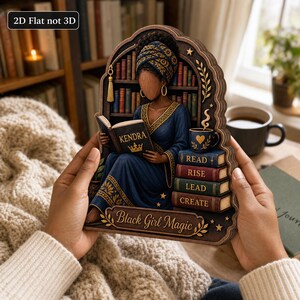 May include: A 2D flat wooden wall art piece featuring a dark-skinned person in a blue dress and headwrap reading a book labeled Kendra. The design includes a bookshelf, a stack of books labeled Read, Rise, Lead, Create, and the text Black Girl Magic.