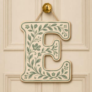 Puede incluir: Una letra E decorativa de madera en color crema con un patrón de enredadera floral pintado a mano en verde salvia, con hojas y pequeñas flores. La letra está suspendida por una cuerda beige sujeta a un gancho metálico dorado.