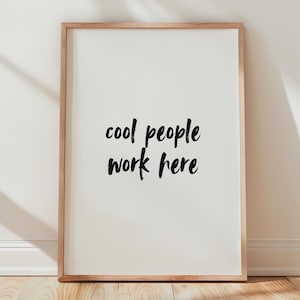 May include: A framed print with the text "cool people work here" in a black, handwritten-style font. The frame is a light brown color, and the print is leaning against a white wall on a wooden floor.