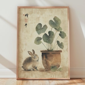 May include: A framed watercolor print featuring a rabbit and a potted plant. The artwork has a beige background with a brown rabbit and a brown pot with green leaves. The frame is made of light brown wood. There is text in the upper left corner.