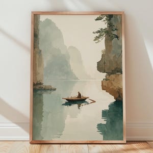 Può includere: Una stampa ad acquerello incorniciata raffigura un paesaggio sereno con una piccola barca su acque calme. L'opera presenta tonalità tenui di blu, verde e beige, con imponenti formazioni rocciose sullo sfondo. La stampa è esposta contro un muro bianco e un pavimento in legno.