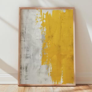 Puede incluir: Impresión artística abstracta en un marco de madera. La obra presenta un diseño texturizado con una división vertical: un lado es gris, el otro es amarillo. La impresión se apoya en una pared blanca sobre un suelo de madera.