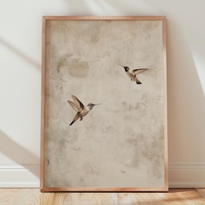 Könnte beinhalten: Ein gerahmter Druck mit zwei Kolibris im Flug vor einem gedämpften, strukturierten Hintergrund. Die Vögel sind detailliert dargestellt und in einem hellbraunen Holzrahmen eingefasst. Das Kunstwerk wird vor einer weißen Wand und einem Holzboden präsentiert.