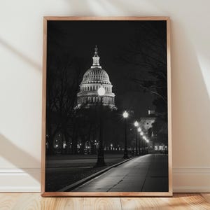 Puede incluir: Fotografía en blanco y negro del Capitolio de los Estados Unidos por la noche, enmarcada en un marco de madera. La imagen captura la cúpula iluminada y los árboles circundantes a lo largo de un camino, creando un contraste dramático.