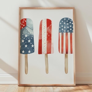 May include: A framed print featuring three watercolor popsicle illustrations. Each popsicle design incorporates red, white, and blue elements reminiscent of the American flag. The popsicles are mounted on wooden sticks, set against a white background.