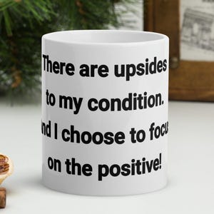 May include: White ceramic mug with the text "There are upsides to my condition, and I choose to focus on the positive!" in black. The mug is next to a dried orange slice and cinnamon sticks.