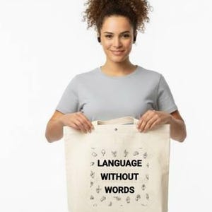 May include: A woman holding a beige tote bag with the text "LANGUAGE WITHOUT WORDS" in black. The bag features small illustrations around the text. The woman is wearing a gray t-shirt and khaki pants.
