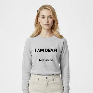 May include: A person wearing a gray sweatshirt and light-colored pants. The sweatshirt has the text "I AM DEAF!" above "Not mute" in black. The person is standing in front of a white background.
