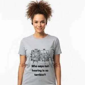 May include: A woman wearing a light gray t-shirt and khaki pants. The t-shirt has a graphic with the text "Who says not hearing is so terrible?!" The woman is standing in front of a white background.