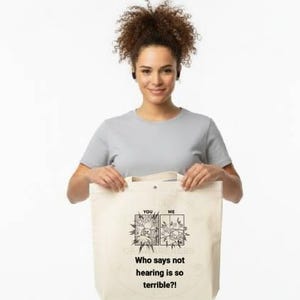 May include: A woman holds a beige tote bag with a black graphic and text that reads, "Who says not hearing is so terrible?!" The bag has two handles and is made of a canvas-like material. The woman is wearing a gray t-shirt and khaki pants.