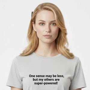 May include: A light gray t-shirt with the text "One sense may be less, but my others are super-powered!" in black. The shirt is worn by a person with blonde hair and is paired with light-colored pants.