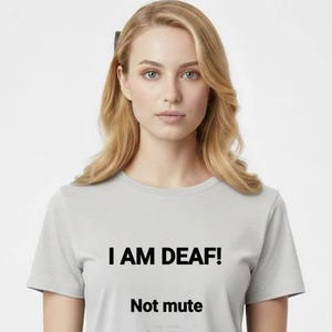 May include: A woman wearing a light gray t-shirt with the text "I AM DEAF!" in black, followed by "Not mute" in a smaller font. The woman is wearing light-colored pants.