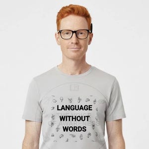 May include: A light gray t-shirt with the text "LANGUAGE WITHOUT WORDS" in black. The shirt features a circular design with various hand sign illustrations. The person in the image is wearing glasses and khaki pants.