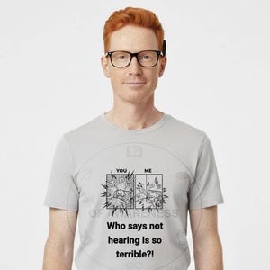 May include: A light gray t-shirt with a graphic that reads "YOU" and "ME" with cartoon images and the text "Who says not hearing is so terrible?!" The person in the image is wearing black glasses and khaki pants.