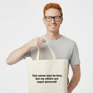 May include: A cream-colored tote bag with the text "One sense may be less, but my others are super-powered!" is held by a person wearing glasses and a gray t-shirt. The bag has a simple design and is held up by the handles.