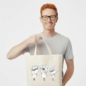 May include: A beige tote bag with the letters "ASL" spelled out in American Sign Language. The bag is held by a person wearing a gray t-shirt and glasses. The background is white.
