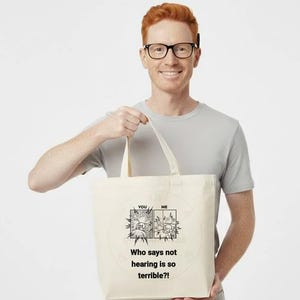 May include: Cream-colored tote bag with a black and white comic strip design and the text "Who says not hearing is so terrible?!" The bag is held by a person wearing glasses and a gray t-shirt.