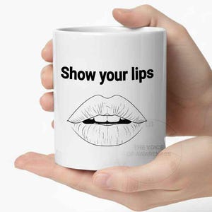 May include: White ceramic mug with the text "Show your lips" in black, above a black and white line drawing of a pair of lips. The mug is held in two hands.