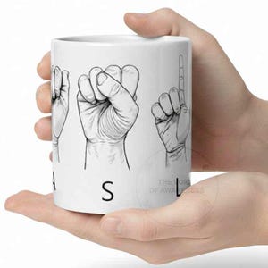 May include: White ceramic mug featuring black line drawings of hand gestures spelling out the word "ASL" in American Sign Language. The mug also has the text "THE VOICE OF AWARENESS".